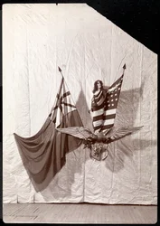 A Living Picture: a model draped in the American Flag standing atop a sculpture of a bald eagle